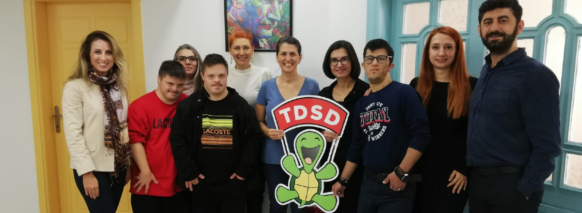 Beneficaries and members of the Türkiye Down Syndrom Association standing for a photo with the charity logo