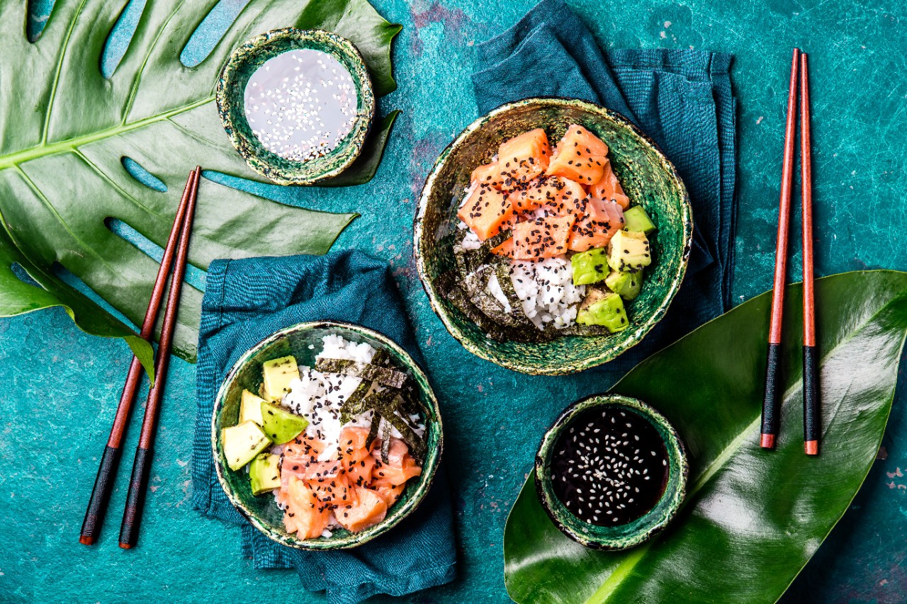 Hawaiian salmon poce with avocado, rice and sesamo served in bowls on tropical leaves. Turquoise slate background. Top view.